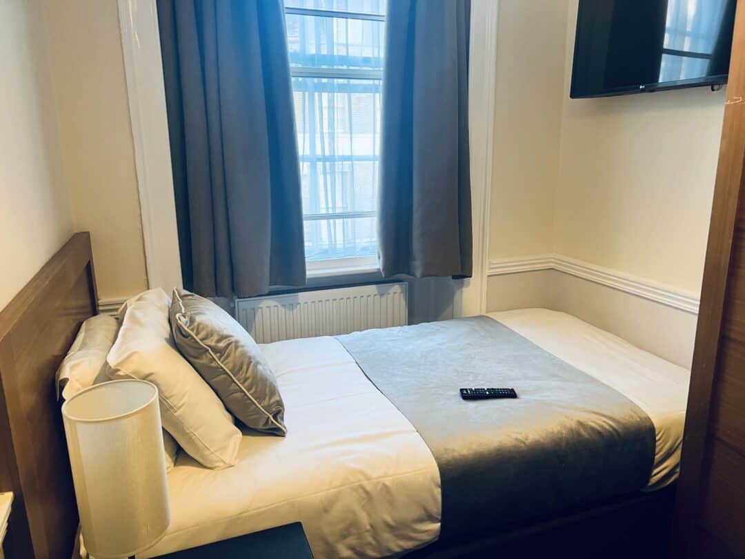 A small, neatly made bedroom with a single bed, gray curtains, a lamp on the nightstand, and a TV mounted on the wall. A remote control rests on the bedspread. Sunlight filters through the window.