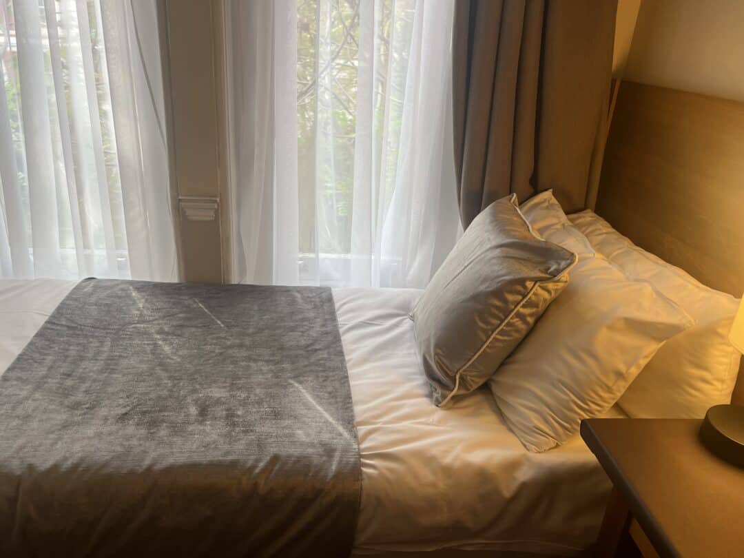A neatly made single bed with white sheets, two gray cushions, and a gray throw blanket, next to a window with sheer curtains and a brown curtain, with sunlight softly illuminating the room.