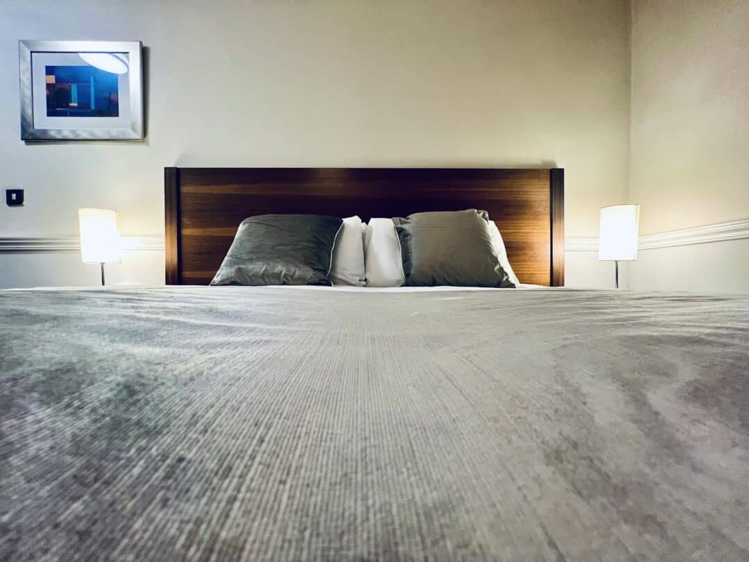 A neatly made bed with two pillows against a wooden headboard, flanked by two lit table lamps. A framed picture hangs on the beige wall above the headboard. The view is from the foot of the bed.