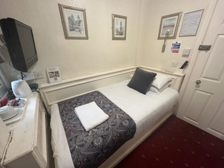 A small hotel room with a single bed, white bedding, a decorative throw and pillow, a folded towel, wall art, a wall-mounted TV, tea-making amenities, and a carpeted red floor.