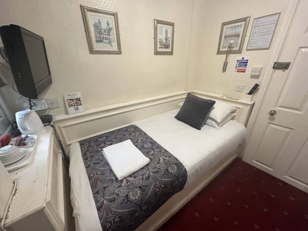 A small hotel room with a single bed, white bedding, a decorative throw and pillow, a folded towel, wall art, a wall-mounted TV, tea-making amenities, and a carpeted red floor.