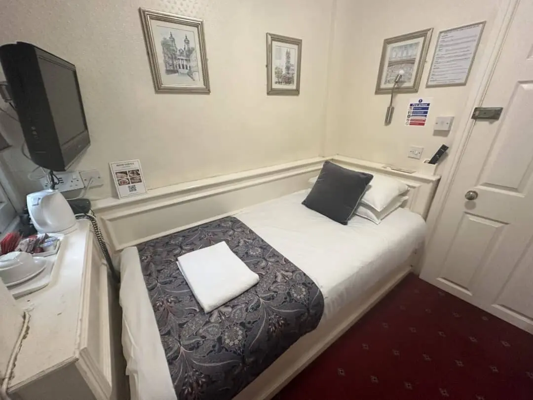 A small hotel room with a single bed, white bedding, a decorative throw and pillow, a folded towel, wall art, a wall-mounted TV, tea-making amenities, and a carpeted red floor.