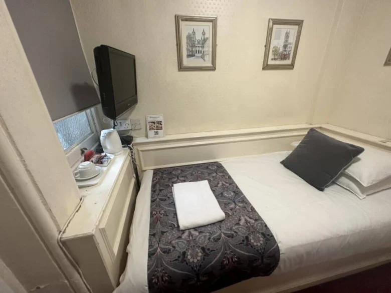 A small, neatly made single bed with a gray pillow and a towel sits by a wall under framed art. A TV is mounted above a windowsill holding a kettle, cups, and tea supplies in a cozy hotel room.