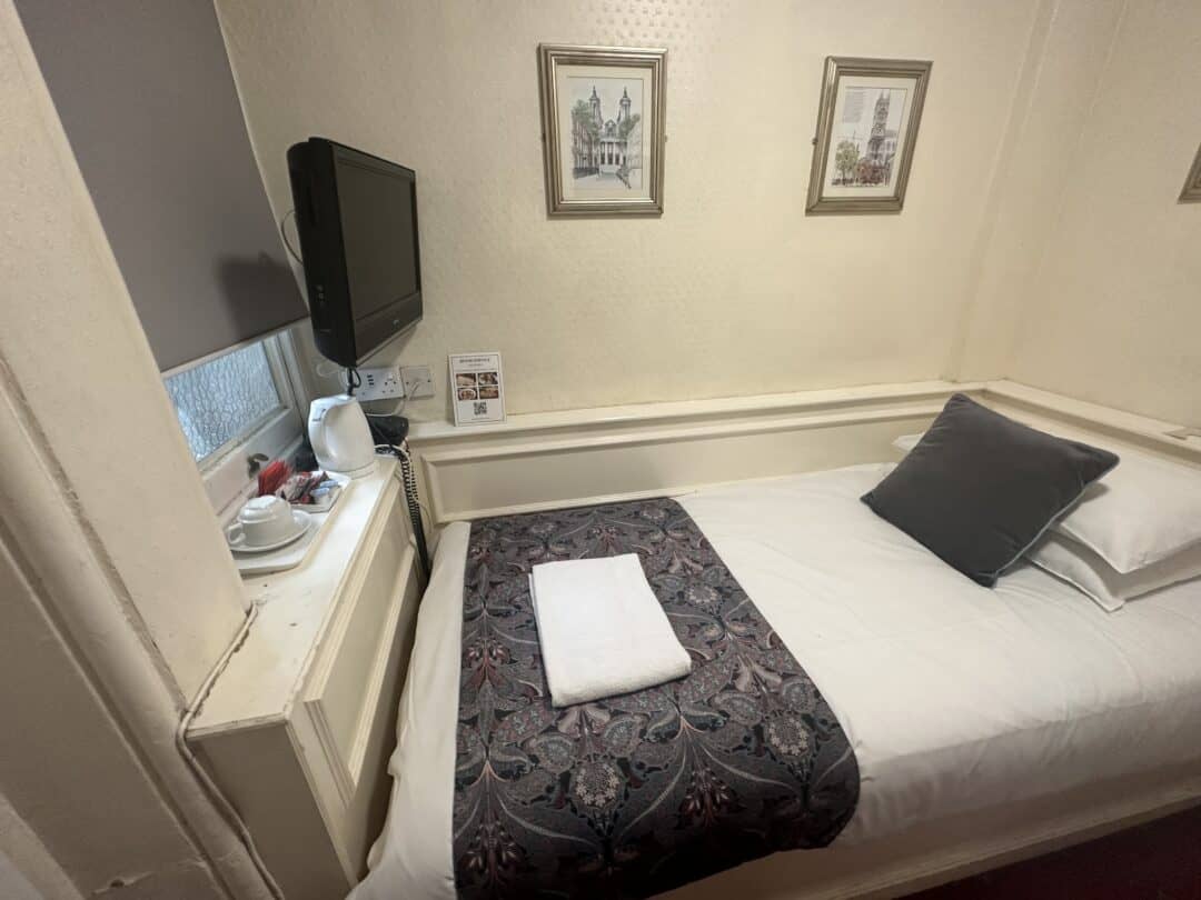 A small, neatly made single bed with a gray pillow and a towel sits by a wall under framed art. A TV is mounted above a windowsill holding a kettle, cups, and tea supplies in a cozy hotel room.