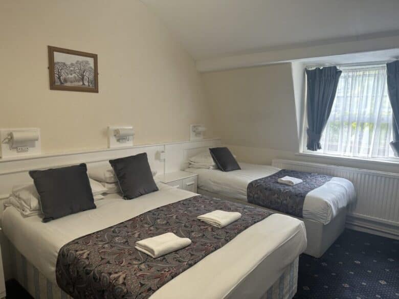 A hotel room with one double bed and one single bed, both neatly made with patterned runners, gray pillows, and folded towels. There’s a window with dark curtains and a framed picture on the cream-colored wall.