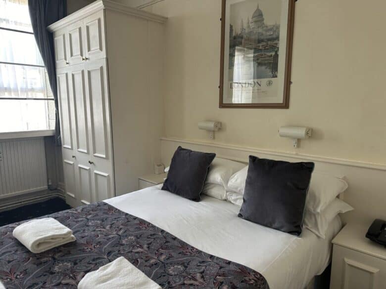 A neatly made double bed with white sheets, two dark gray pillows, and a patterned runner, sits in a bright room with a large wardrobe, a framed London print, and two folded towels at the foot of the bed.