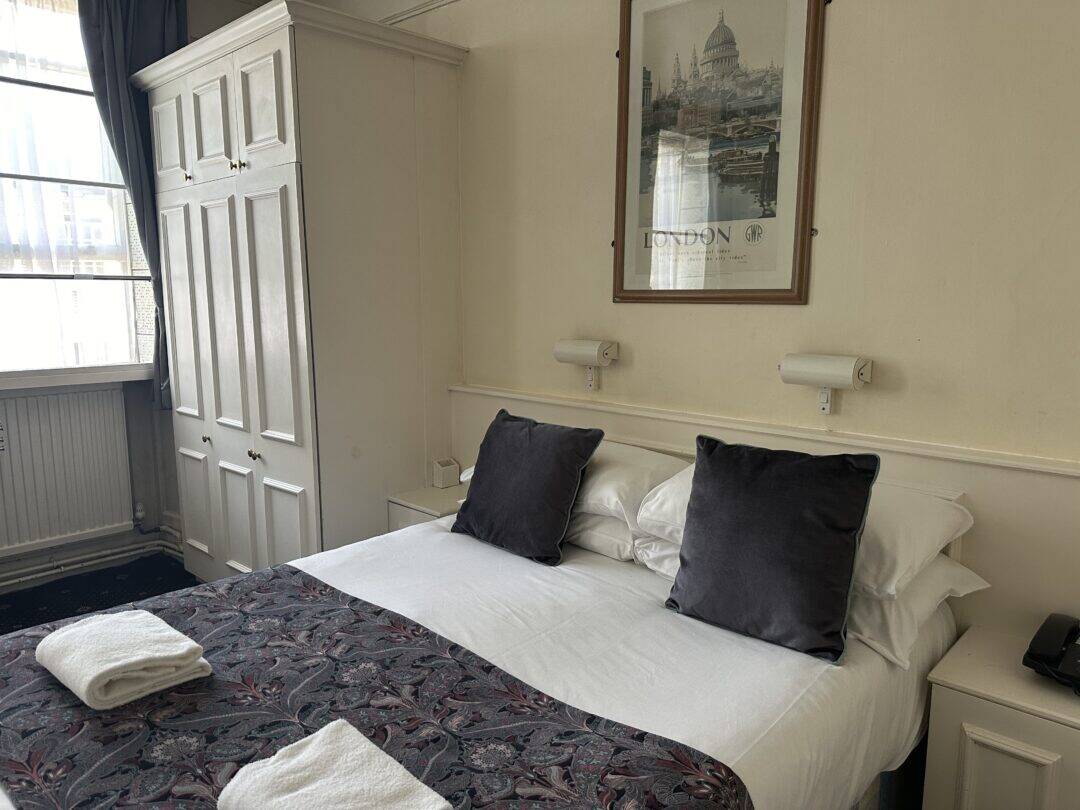 A neatly made double bed with white sheets, two dark gray pillows, and a patterned runner, sits in a bright room with a large wardrobe, a framed London print, and two folded towels at the foot of the bed.