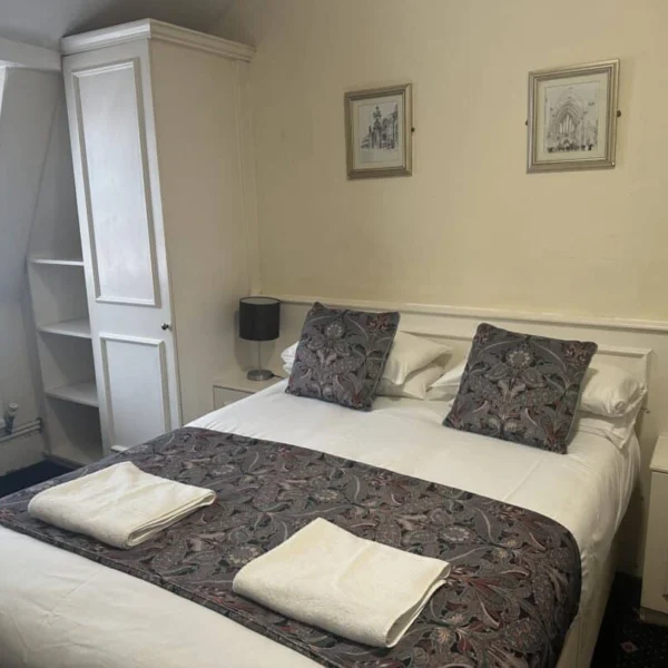 A neatly made double bed with two patterned pillows and matching runner, two folded white towels, flanked by a lamp and phone on bedside tables, in a cozy, softly lit hotel room with white furniture and framed art above the bed.