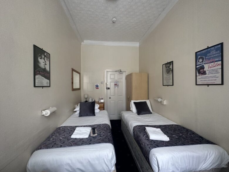A small hotel room with two single beds, each with a folded towel and a dark runner. The walls are beige, decorated with framed posters. A wardrobe and mirror are near the white door at the end of the room.