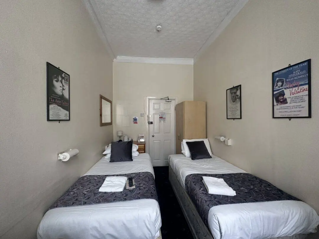 A small hotel room with two single beds, each with a folded towel and a dark runner. The walls are beige, decorated with framed posters. A wardrobe and mirror are near the white door at the end of the room.