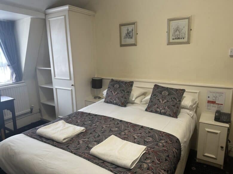 A neatly made double bed with two patterned pillows and matching runner, two folded white towels, flanked by a lamp and phone on bedside tables, in a cozy, softly lit hotel room with white furniture and framed art above the bed.