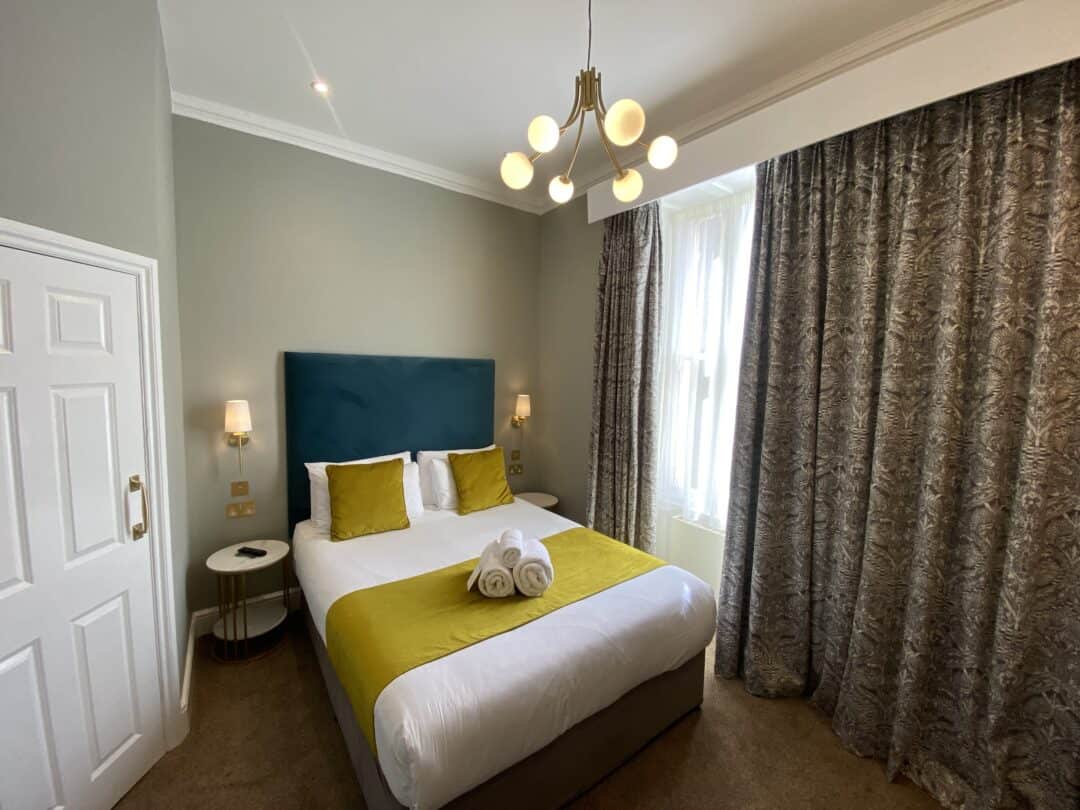 A modern hotel room with a double bed, teal headboard, yellow and white bedding, two bedside tables with lamps, patterned curtains, and a window letting in natural light. Towels are neatly rolled on the bed.