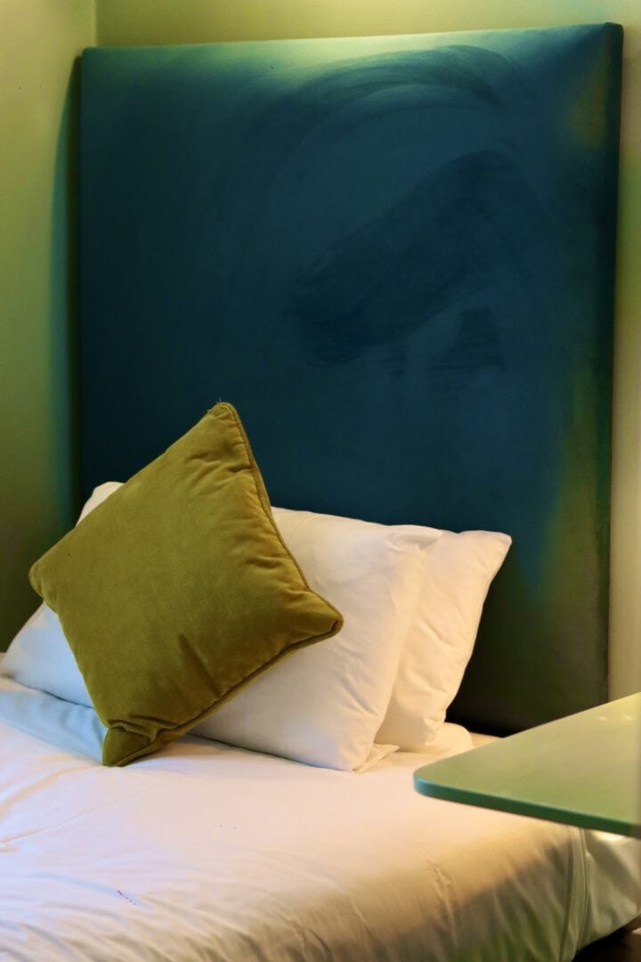 A neatly made bed with white sheets and two white pillows, topped with a green cushion, sits against a tall, teal upholstered headboard in a softly lit room.