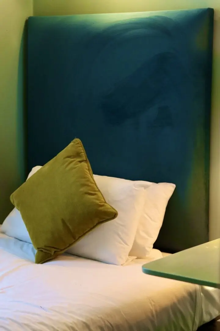 A neatly made bed with white sheets and two white pillows, topped with a green cushion, sits against a tall, teal upholstered headboard in a softly lit room.