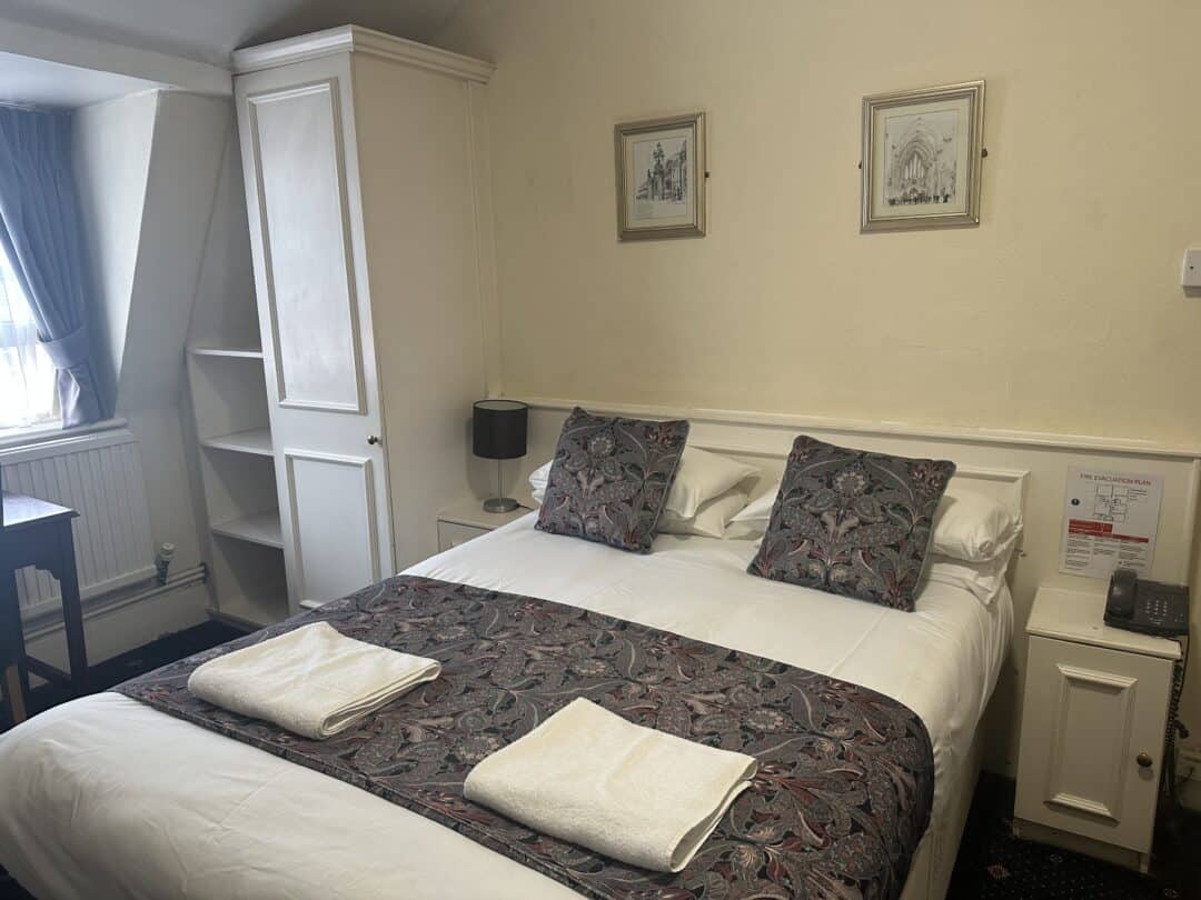 A neatly made double bed with two patterned pillows and matching runner, two folded white towels, flanked by a lamp and phone on bedside tables, in a cozy, softly lit hotel room with white furniture and framed art above the bed.