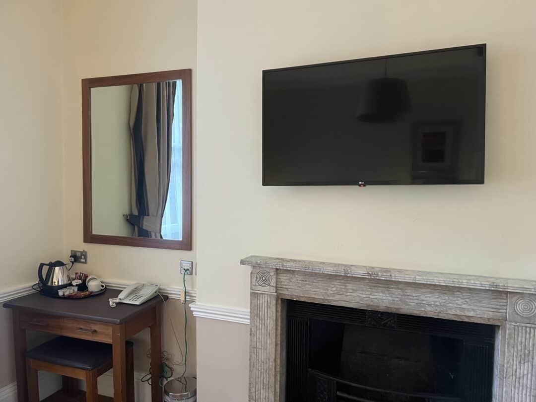 A wall-mounted TV above a fireplace, a mirror reflecting a window with curtains, a wooden desk with a kettle and mugs, a telephone on the wall, and a waste bin underneath the desk in a light-colored room.