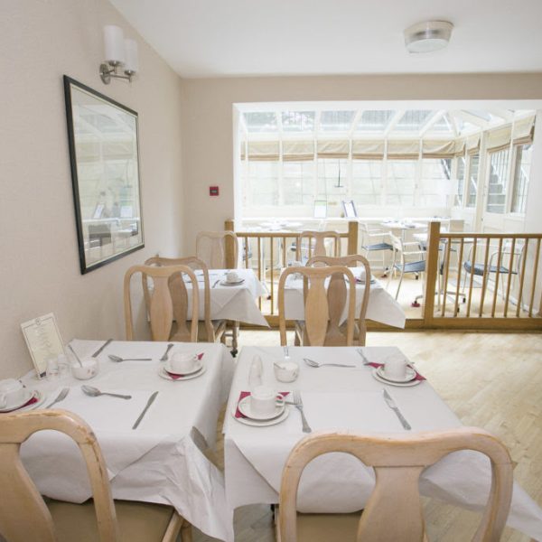 A bright dining room in one of the charming hotels in London features light wood flooring and white walls. It has four tables, each set with white tablecloths, dishes, cups, and cutlery. Large windows and an adjoining sunlit area with additional seating can be seen in the background.