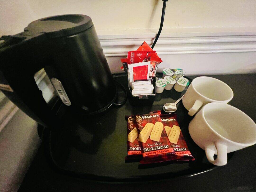 A black electric kettle, two white mugs, creamers, sugar packets, tea and coffee sachets, and three packs of Walkers shortbread cookies are arranged on a black counter.