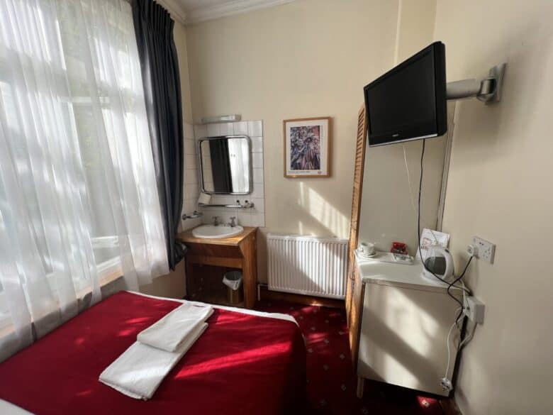 A small hotel room with a single bed covered in a red blanket, a towel folded on top, a wall-mounted TV, a sink with a mirror, a desk with a kettle, and a window with sheer white curtains.