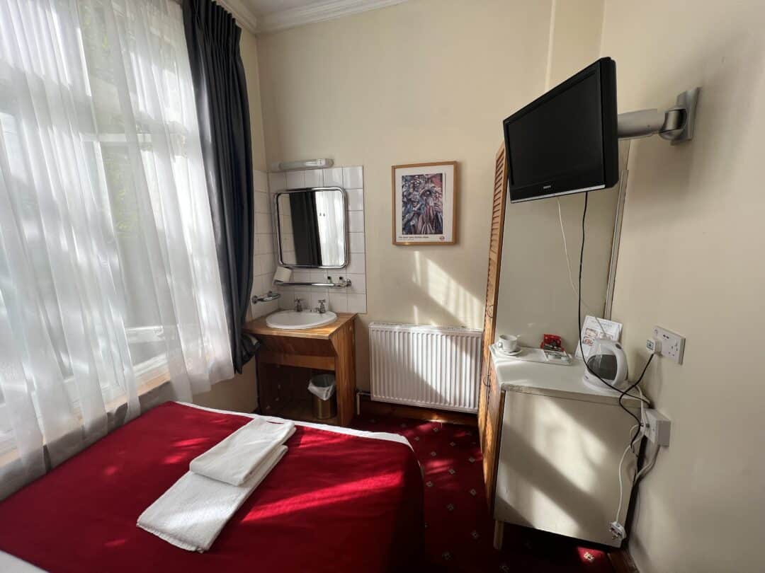 A small hotel room with a single bed covered in a red blanket, a towel folded on top, a wall-mounted TV, a sink with a mirror, a desk with a kettle, and a window with sheer white curtains.
