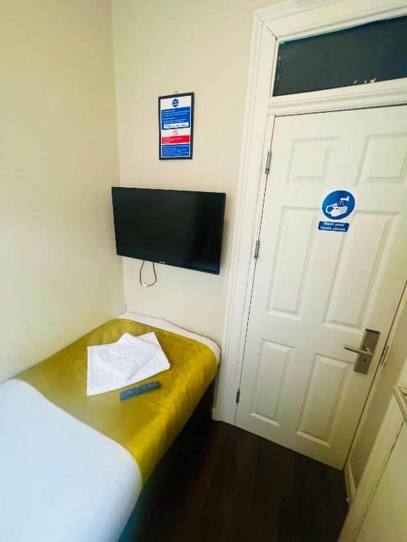 A small, tidy room with a single bed, a yellow blanket, a folded towel, and a TV mounted on the wall. A white door with a blue “fire door keep shut” sign and a remote control are also visible.