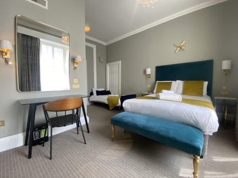 A modern hotel room with a double bed featuring green velvet headboard, yellow and white bedding, a teal bench, desk with chair, large mirror, wall lamps, and a window with white curtains.