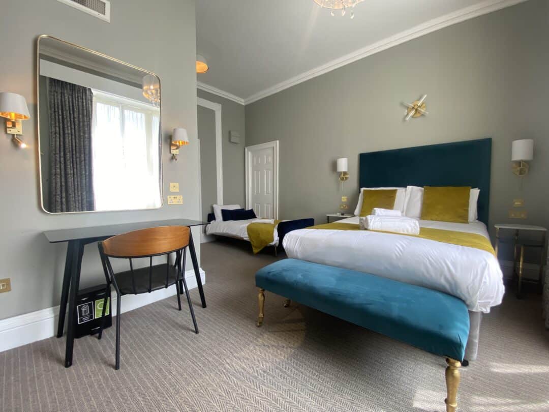 A modern hotel room with a double bed featuring green velvet headboard, yellow and white bedding, a teal bench, desk with chair, large mirror, wall lamps, and a window with white curtains.