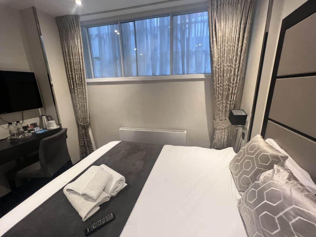 A neatly made hotel room bed with two folded towels and a remote on top. A desk with a chair, TV, kettle, and cups are on the left. Light curtains frame a large window above the bed.