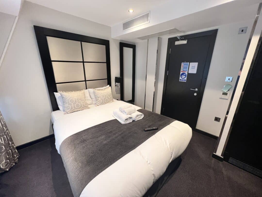 A modern hotel room with a neatly made double bed, two patterned pillows, folded towels, and a TV remote on a gray blanket. The room features dark furniture, a door, and a wall-mounted mirror.