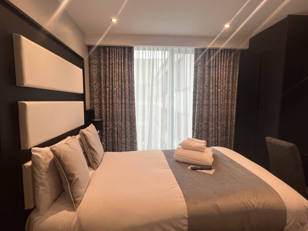 A neatly made bed with gray and white bedding sits in a modern hotel room, with folded towels and a TV remote placed on top. The room has dark walls, patterned curtains, and large windows letting in natural light.