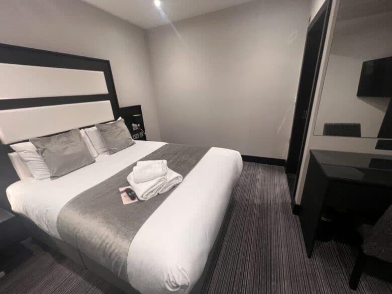 A modern hotel room with a double bed covered in white and gray linens, three pillows, and folded towels on top. There is a desk and chair with a mirror, and the walls are painted a light color.