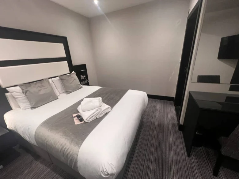 A modern hotel room with a double bed covered in white and gray linens, three pillows, and folded towels on top. There is a desk and chair with a mirror, and the walls are painted a light color.