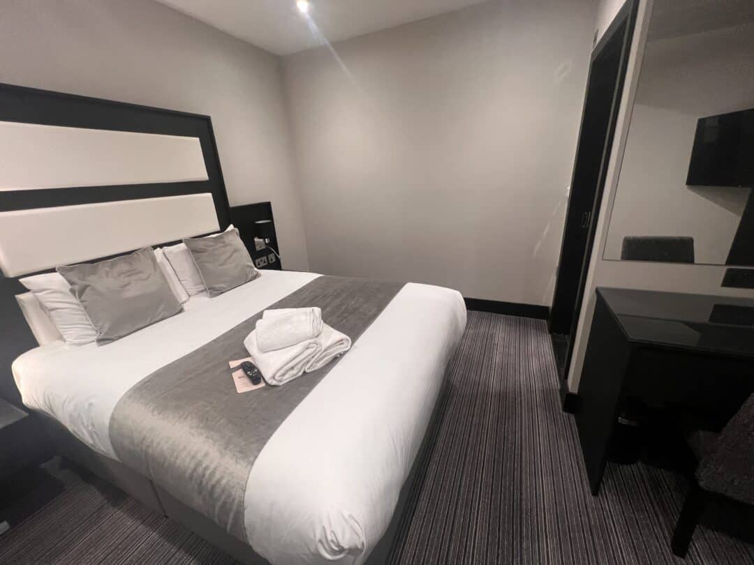 A modern hotel room with a double bed covered in white and gray linens, three pillows, and folded towels on top. There is a desk and chair with a mirror, and the walls are painted a light color.