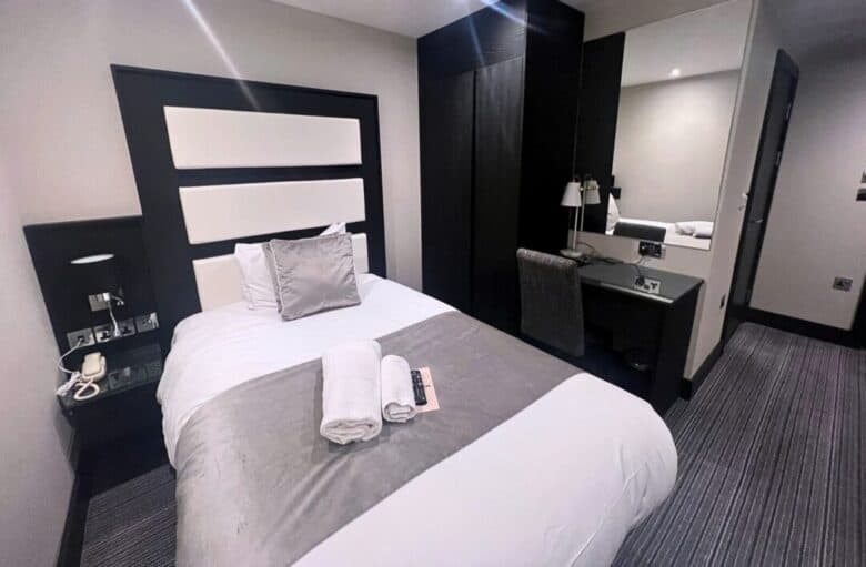A modern hotel room with a single bed, gray and white bedding, a headboard, a side table with a phone, and a desk with a mirror, lamp, chair, and TV remote. Two towels are neatly placed on the bed.