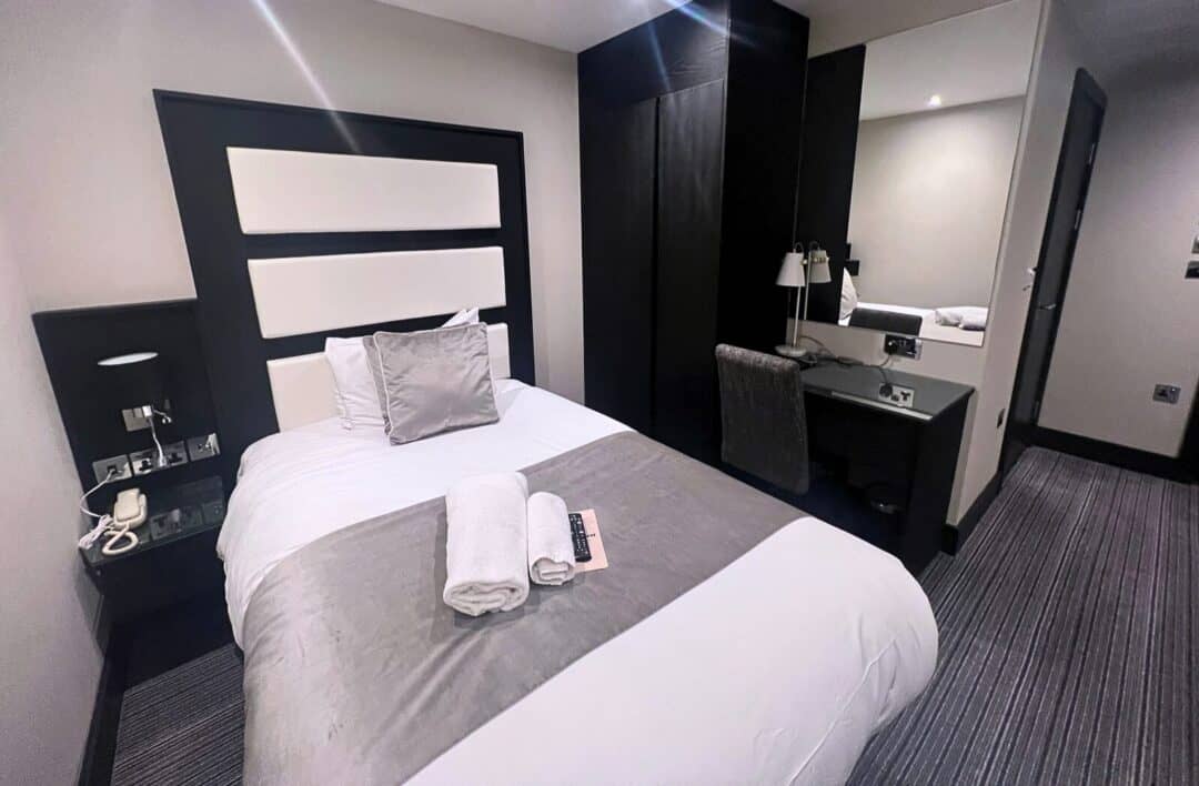 A modern hotel room with a single bed, gray and white bedding, a headboard, a side table with a phone, and a desk with a mirror, lamp, chair, and TV remote. Two towels are neatly placed on the bed.