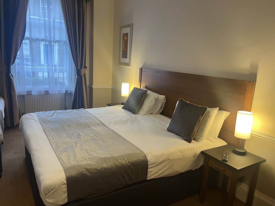 A neatly made bed with gray and white bedding sits between two nightstands with lit lamps in a cozy, softly lit hotel room. A large window with curtains is on the left wall.
