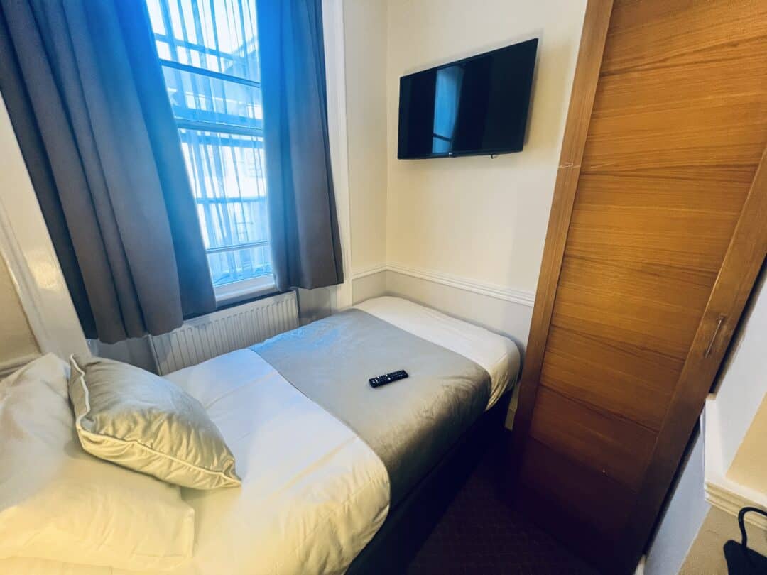 A small hotel room with a single bed, a pillow, a folded blanket, a TV mounted on the wall, a wardrobe, a window with curtains, and a TV remote on the bed.