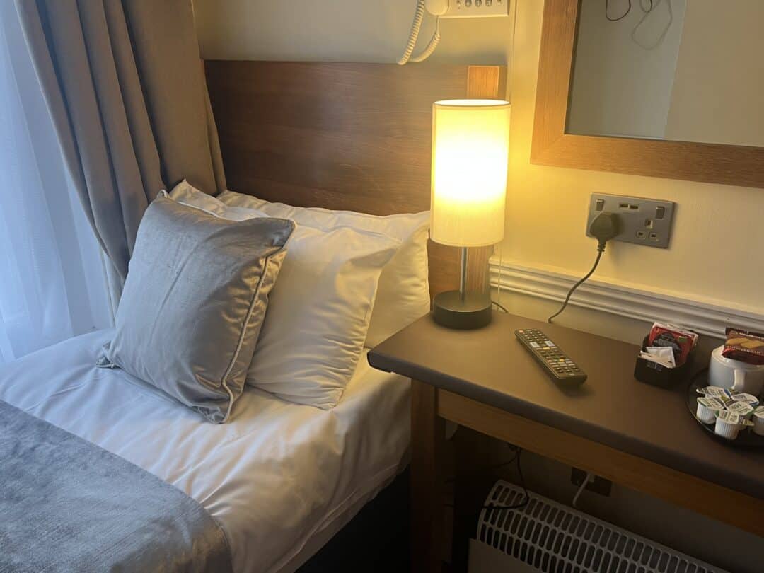 A neatly made bed with white and grey pillows next to a wooden nightstand holding a lit lamp, TV remote, snacks, and tea supplies. A large window with sheer curtains is on the left.