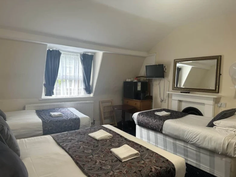 A cozy hotel room with three single beds, each topped with a folded towel and patterned runner. There's a window with dark curtains, a desk with a TV and mini fridge, a chair, and a large mirror above a fireplace.