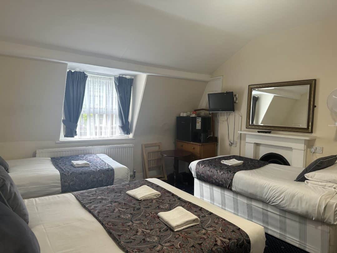 A cozy hotel room with three single beds, each topped with a folded towel and patterned runner. There's a window with dark curtains, a desk with a TV and mini fridge, a chair, and a large mirror above a fireplace.