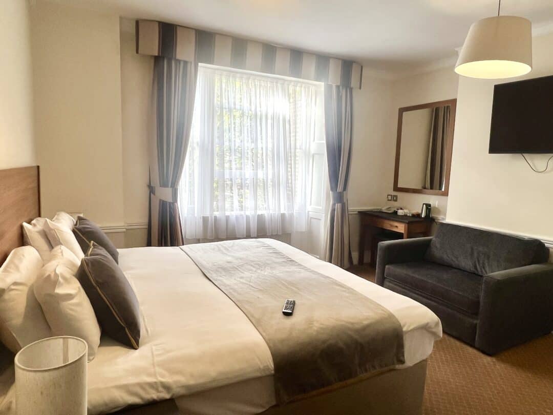 A bright hotel room with a double bed, striped curtains, sofa, wall-mounted TV, desk with mirror, and a lamp. A TV remote rests on the neatly made bed under a large window letting in natural light.