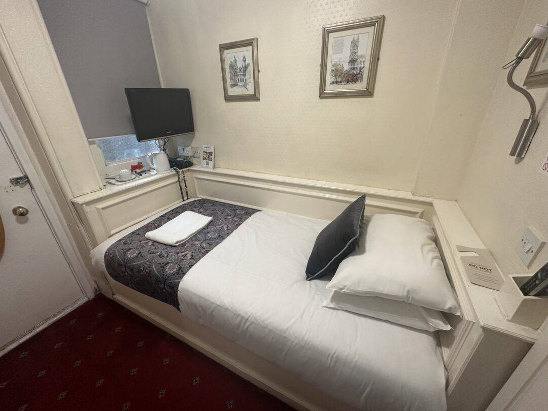A small hotel room with a single bed, white sheets, and a dark patterned runner. There are two pillows, a folded towel, a wall-mounted TV, a tea set, two framed pictures, and a wall lamp. The carpet is red.
