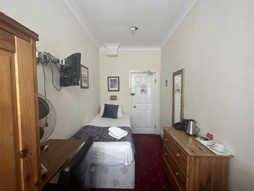 A narrow hotel room with a single bed, wooden wardrobe, dresser, wall-mounted TV, desk fan, mirror, and tea-making supplies. The room has cream walls and a red carpet with a white door at the end.
