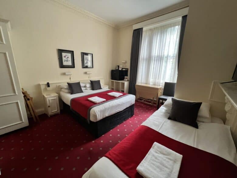 A hotel room with two beds, each made with white linens and red throws, a small desk and chair by a large window with sheer curtains, and framed artwork on the beige walls. A folded towel is on each bed.