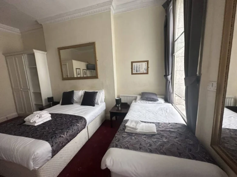 A hotel room with two beds, one double and one single, both with white bedding and dark patterned runners. A folded towel is on each bed. There are large mirrors, a window with gray curtains, and framed art on cream-colored walls.