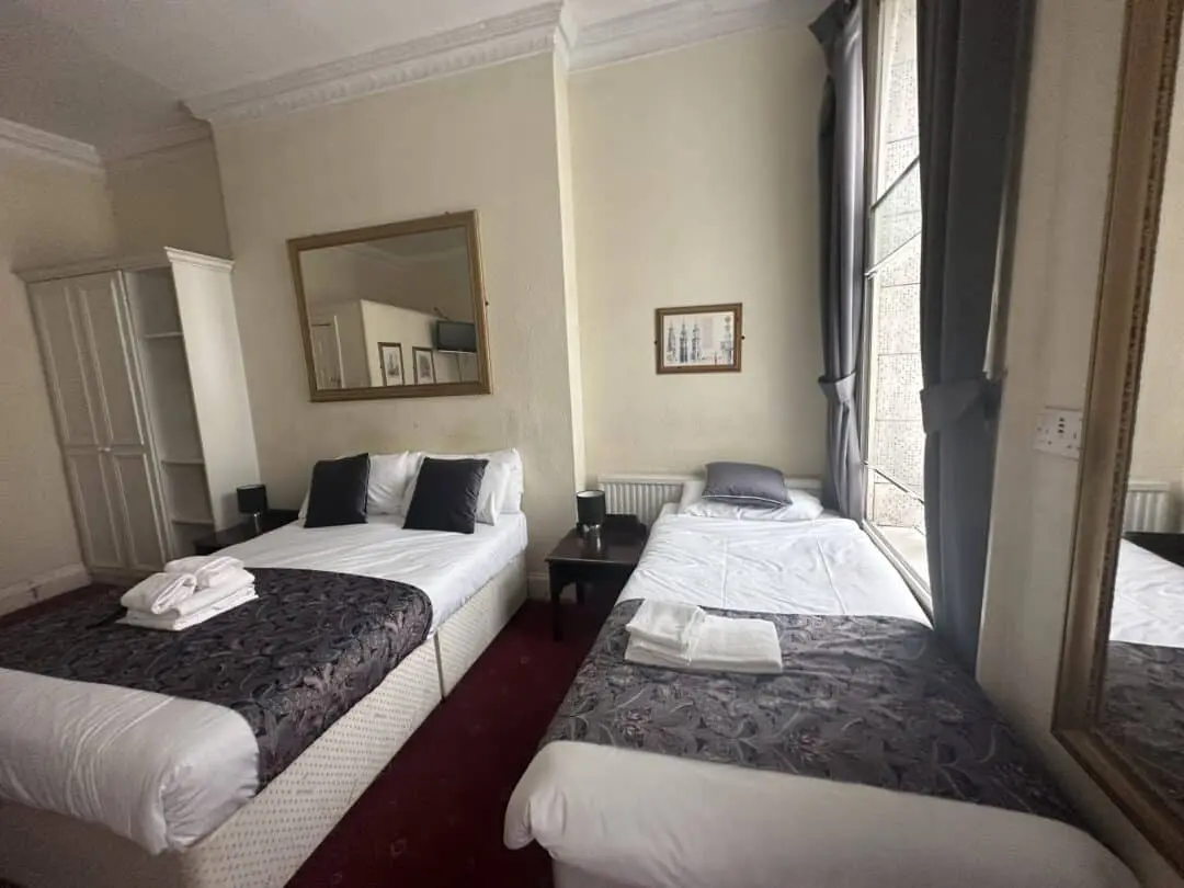 A hotel room with two beds, one double and one single, both with white bedding and dark patterned runners. A folded towel is on each bed. There are large mirrors, a window with gray curtains, and framed art on cream-colored walls.
