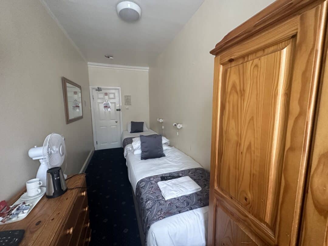A narrow hotel room with a single bed, two gray pillows, a folded towel, a wooden wardrobe, a dresser with a fan, a kettle, and cups, and a door at the far end. The walls are cream-colored.