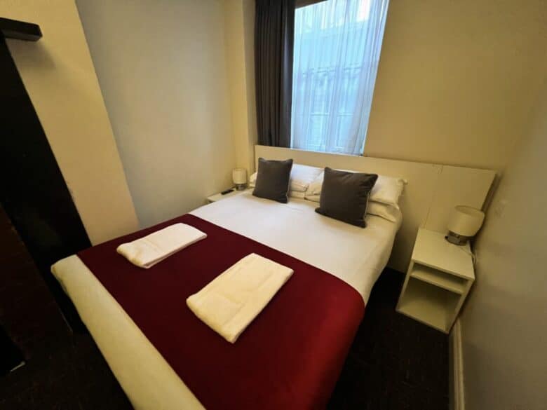 A small, simply furnished bedroom with a double bed, white bedding, a red throw, two folded white towels, two dark pillows, bedside tables with lamps, and a window with sheer curtains letting in daylight.