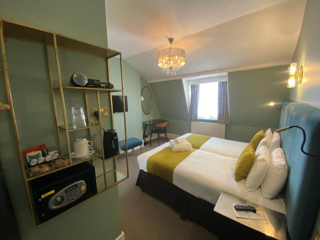 A cozy hotel room with a double bed, green walls, a wall-mounted open shelf with a kettle, cups, and a safe, a chandelier, a window with patterned curtains, and a small round mirror above a blue stool.
