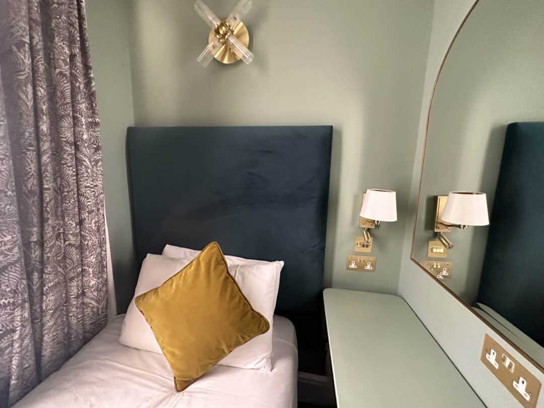 A neatly made bed with a green upholstered headboard, a white pillow, and a yellow decorative cushion sits beside a wall-mounted lamp, floral curtains, and a vanity with a large mirror and two small wall lamps.